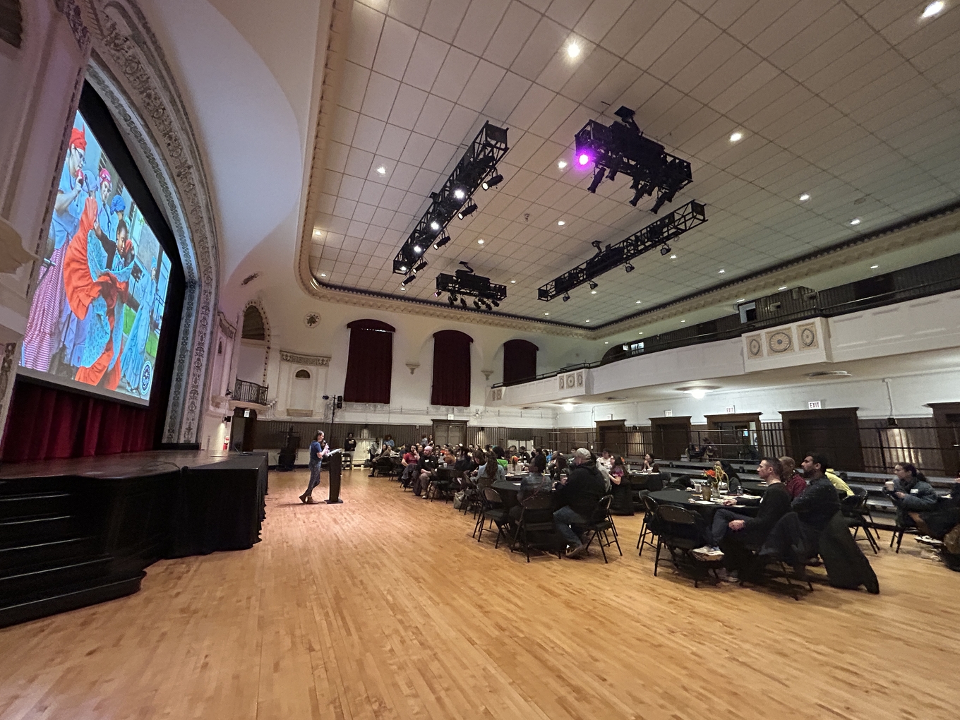 A speaker presents to an audience seated at round tables in a large auditorium. A projected image of people in colorful clothing is visible on a screen behind the stage.